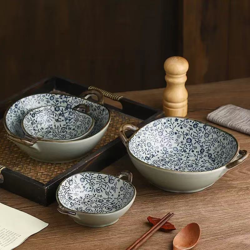 Three ceramic bowls with blue floral patterns and brown handles, arranged on a wooden table with a tray, pepper mill, cloth, chopsticks, and a spoon.
