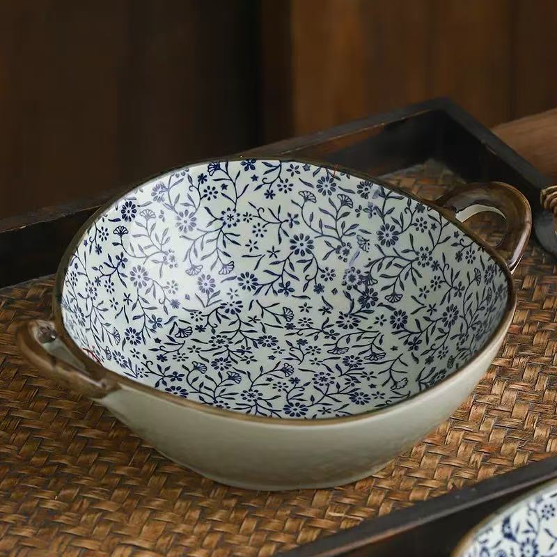A ceramic bowl with a blue floral pattern on a white interior and a beige exterior, featuring two brown handles, displayed on a woven mat.