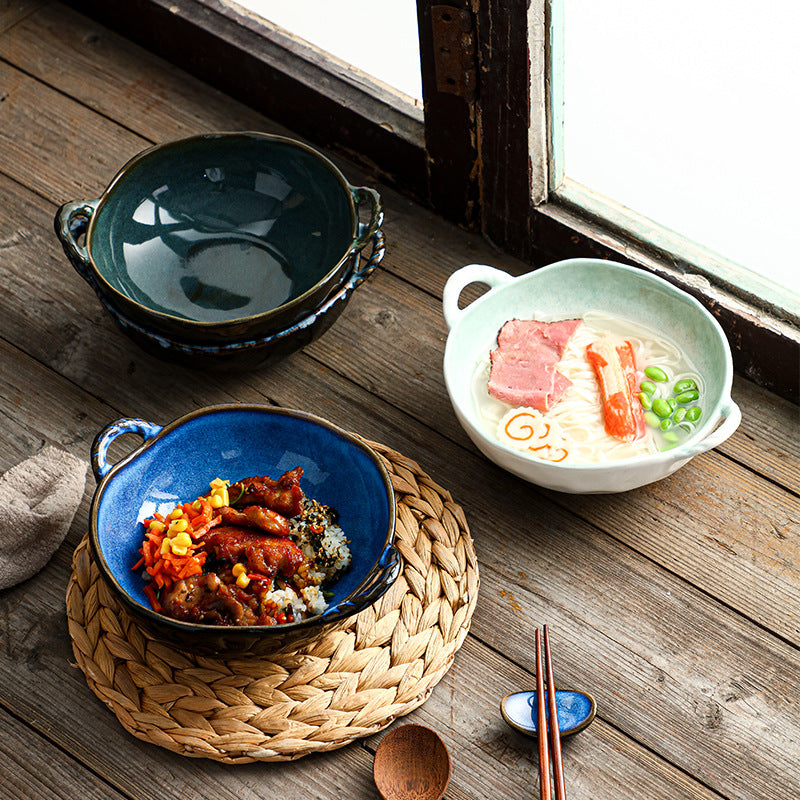 A styled tabletop scene with ceramic bowls and a bowl of ramen on a rustic wooden table near a window. The set includes a dark teal bowl, a blue bowl with food, a white bowl with ramen, chopsticks, a spoon, and a woven placemat.