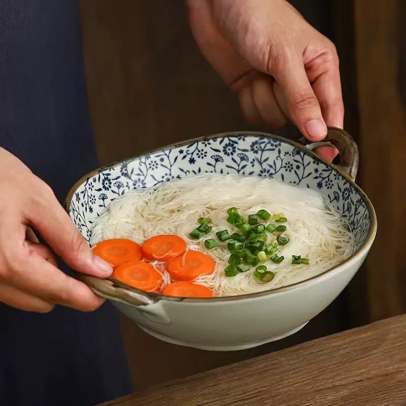 A person holds a ceramic bowl of noodle soup with thin white noodles, sliced orange carrots, and chopped green onions. The bowl has a blue floral pattern on the inside, a beige exterior, and dark brown handles, set against a dark wooden background.