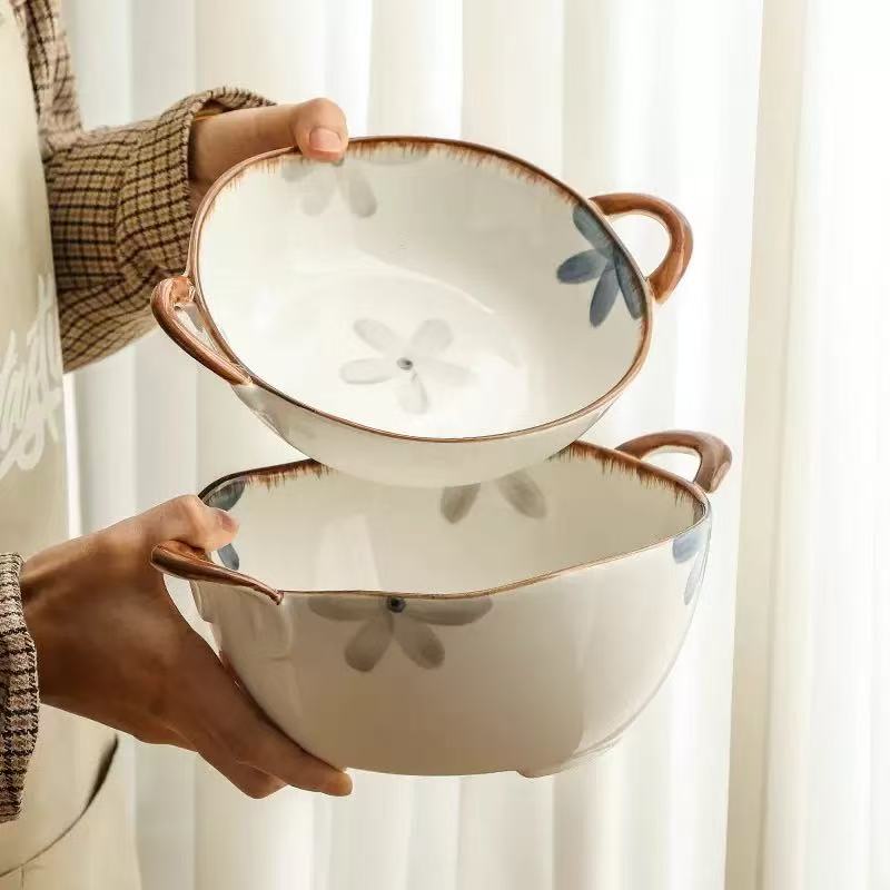 Two ceramic bowls with handles, held by a person. The bowls are off-white with a brown rim, featuring a central floral motif in gray and blue.