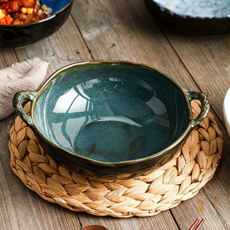 A dark teal glazed ceramic bowl with side handles sits on a woven straw placemat on a rustic wooden table. In the background, other bowls and dining props are visible.