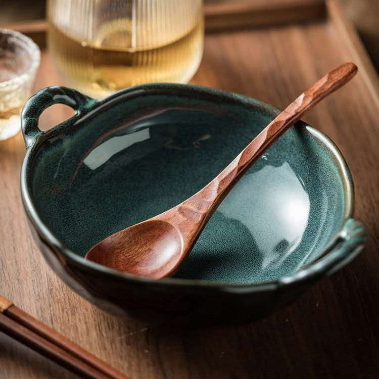 A dark teal ceramic bowl with side handles and a wooden spoon resting inside, set on a wooden tray with a glass of tea in the background.