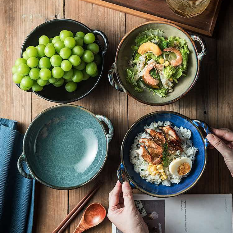 Four ceramic bowls with handles are arranged on a wooden table, styled with food and dining accessories. One bowl is filled with green grapes, one with a salad, one is empty, and one is being held by hands over rice with grilled chicken and a hard-boiled egg.
