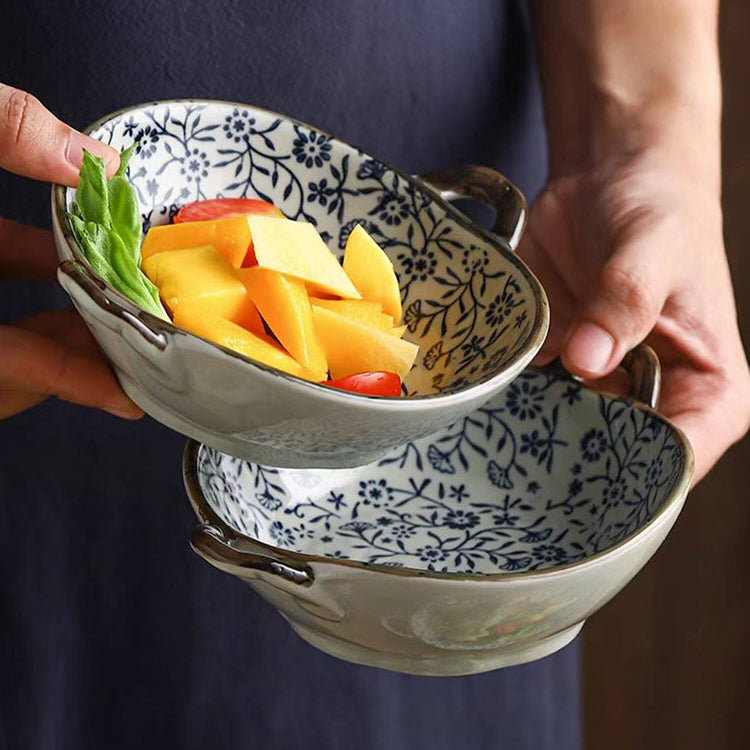 A person in a navy apron holds two ceramic bowls with a blue floral pattern and dark handles, one bowl containing sliced mangoes and green leaves.