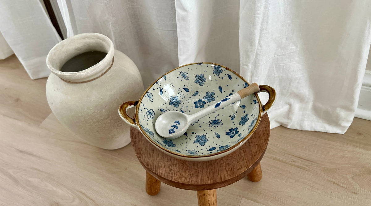 A ceramic bowl with blue floral patterns and gold handles sits on a small wooden stand, with a matching spoon resting inside. The setup is placed on a light wood floor in front of white curtains, with a large beige ceramic vase in the background.