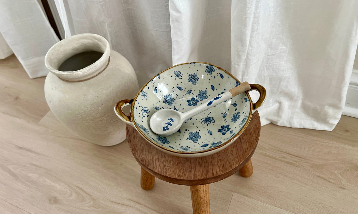 A ceramic bowl with blue floral patterns and gold handles sits on a small wooden stand, with a matching spoon resting inside. The setup is placed on a light wood floor in front of white curtains, with a large beige ceramic vase in the background.