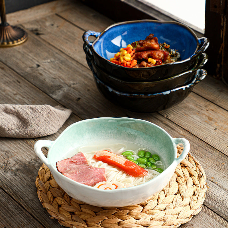 A ceramic bowl of noodle soup with ham, crab stick, green beans, and rice sits on a woven placemat on a rustic wooden table, with a stack of dark blue and black bowls in the background.