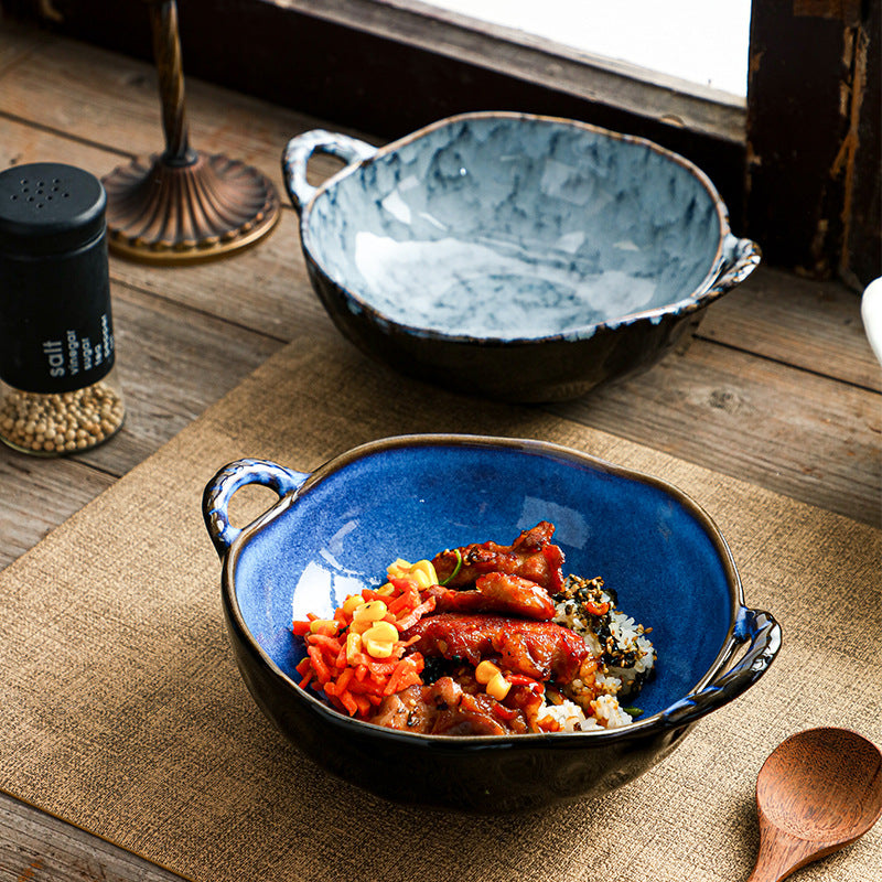 A rustic ceramic bowl with a deep blue interior and dark exterior, filled with rice and colorful food, set on a wooden table with a second empty bowl in the background.