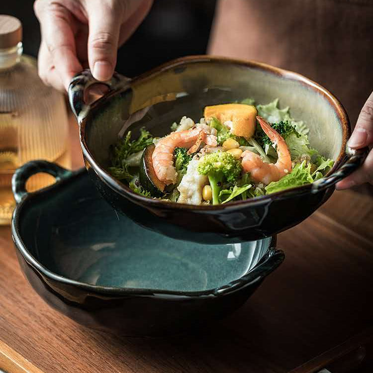 A person holds a dark ceramic bowl filled with a shrimp salad over another empty matching bowl, set on a wooden table.