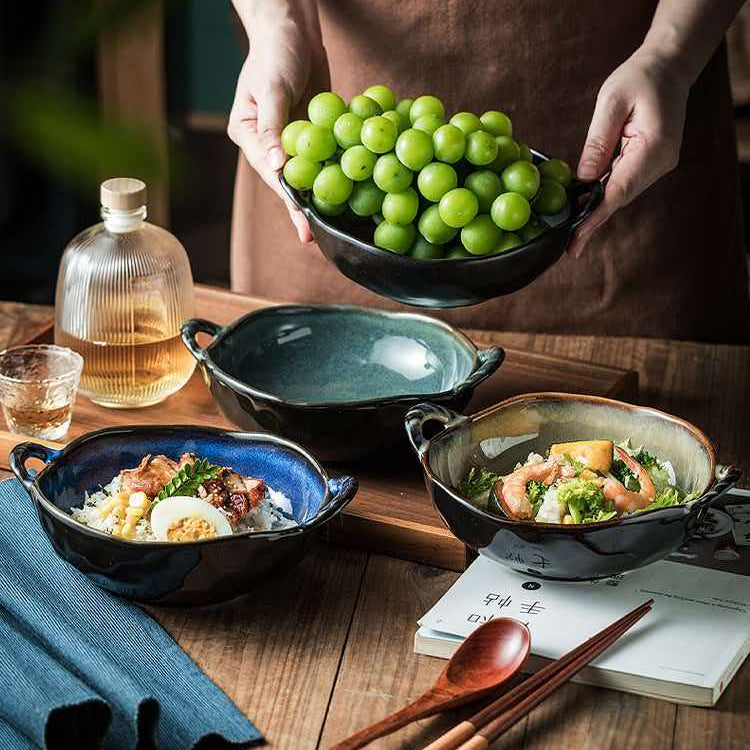 A person in a brown apron holds a black bowl filled with green grapes over a rustic wooden table. Several ceramic bowls with food, a glass bottle, chopsticks, a spoon, and a notebook are arranged on the table.
